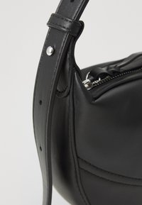 Μαύρη δερμάτινη τσάντα με καμπυλωτό σχήμα, διαθέτοντας υφασμάτινη επιφάνεια, ρυθμιζόμενο λουρί και ασημένια μεταλλικά στοιχεία. Ορατή λεπτομέρεια φερμουάρ.