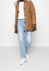 Parka marrone con cappuccio foderato in pelliccia, chiusura frontale con zip e bottoni, abbinata a jeans azzurri e sneakers bianche. Materiali lisci e vestibilità casual.