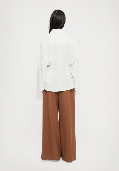 White blouse with long sleeves, tailored fit, and a back seam detail paired with wide-legged brown trousers in a smooth fabric.