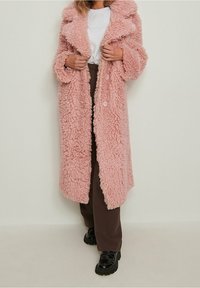 Abrigo largo, esponjoso y rosa con un exterior texturizado, gran cuello y dos botones, combinado con pantalones marrones y zapatos negros.