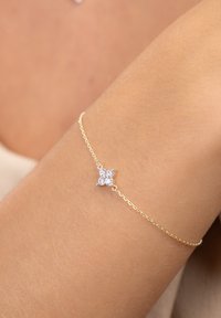 Goldene Kettenarmband mit floraler Gestaltung und drei klaren Edelsteinen in der Mitte, auf einem gebräunten Hautton.