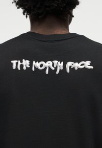 Άτομο με μαύρο πουκάμισο που φέρει λευκό στυλιζαρισμένο κείμενο "THE NORTH FACE" στην κορυφή της πλάτης, φωτογραφημένο από πίσω με απλό φόντο.