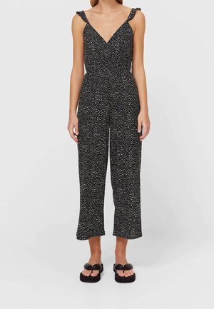 Zwarte jumpsuit met witte stippen, v-hals, gerimpelde schouderbanden en cropped pijpen. Gestyled met zwarte sandalen met strikaccenten.