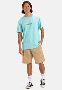 Camiseta de algodón turquesa con el logotipo de Quiksilver, combinada con pantalones cortos cargo beige. El modelo lleva zapatillas negras y calcetines blancos a rayas.