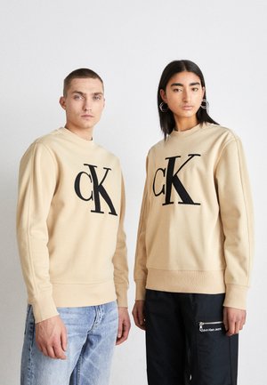 Sudaderas beige con un gran logotipo negro "CK", con puños acanalados y cuello redondo. Combinadas con jeans de color azul claro y pantalones oscuros.