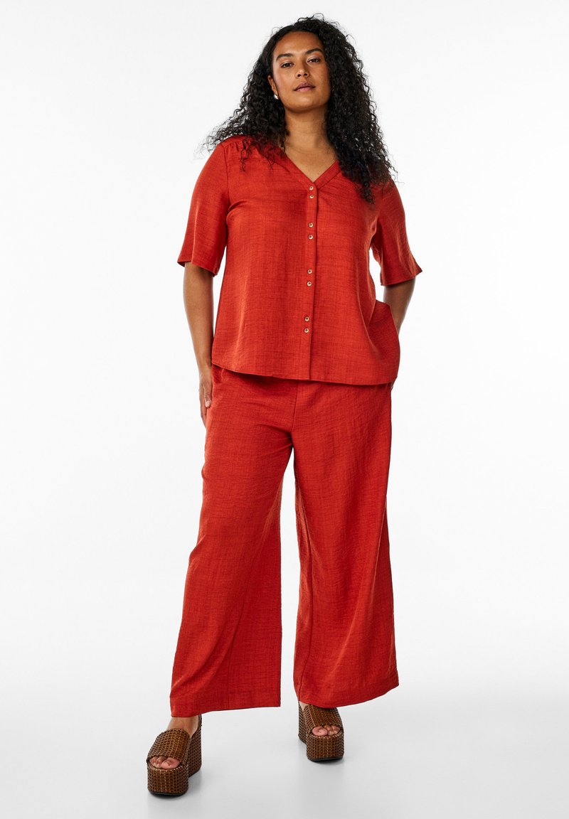 Red, textured linen top with short sleeves and button closure, paired with wide-leg pants; worn with chunky, woven platform shoes.