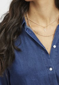 Modrá džínová košile s límcem na knoflíky, doplněná o vrstvené zlaté náhrdelníky s křížkem a malými dekorativními prvky.