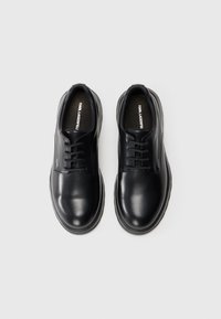 Chaussures en cuir noir avec un bout arrondi, un design à lacets, une texture lisse et une semelle en caoutchouc basse. Logo de la marque visible sur le côté et sur la semelle intérieure.