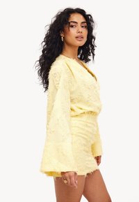 Haut jaune texturé avec des manches à cloche associé à une jupe mini texturée coordonnée. Le tissu semble doux avec des bords effilochés et un léger éclat.