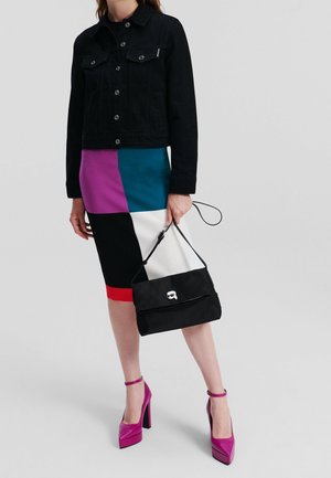 Vrouw draagt een zwart jasje, colorblock-rok, roze hoge hakken, houdt een zwarte handtas met polsriem vast.