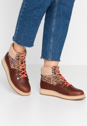 Bottes marron et beige à la cheville avec des lacets orange, tissu à imprimé tigre, bord en polaire, portées avec un jean bleu raccourci sur un sol blanc.