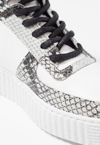 Vita sneakers med en texturerad ormtrycksdesign i grått. Svarta skosnören och en mönstrad vit sula. Detaljerad sömnad synlig.