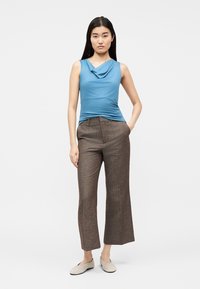 Vrouw staand met hand in de zak, draagend een mouwloze blauwe top, brede bruine broekspijpen en beige platte schoenen tegen een effen witte achtergrond.