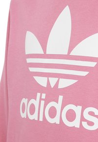 Logo y texto blanco de Adidas Trefoil sobre tela rosa, mostrando parte de un sudadera o hoodie.