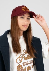 Maroonfarbene Baseballmütze mit gelber, gestickter Schrift; strukturierte Krone, gebogener Schirm und verstellbarer Riemen auf der Rückseite; getragen von einem Model.