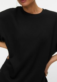 Ženska v preprosti črni majici s kratkimi rokavi, ena roka na boku, na prstu viden srebrn prstan in nežna viseča uhani.