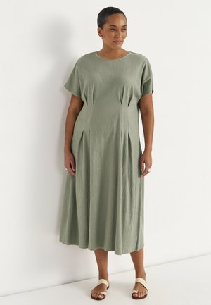 Vrouw draagt een saliegroene gestructureerde midi-jurk met korte mouwen en naaddetails, gecombineerd met beige sandalen, staand tegen een witte muur.