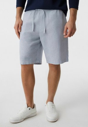 Mężczyzna w jasnoszortach z troczkami, białych trampkach i czarnym topie z długim rękawem, stojący na jednolitym białym tle.