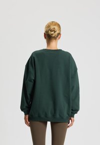 Felpa oversized verde scuro con spalle scese e polsini a costine, realizzata in tessuto morbido. Cuciture visibili e scollatura a girocollo.