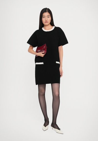 Woman standing in black short-sleeve dress with white trim, sheer black tights, white heels, and holding a red clutch bag.