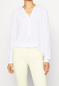 Camicia bianca a maniche lunghe con scollo henley, caratterizzata da una patta di bottoni e una vestibilità leggermente rilassata, abbinata a pantaloni giallo chiaro.