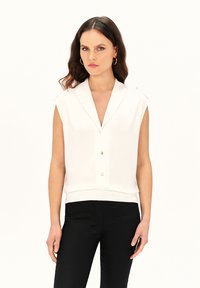 Weißes ärmelloses Oberteil mit einem eingekerbten Kragen, Knöpfen vorne und einem taillierten Saum. Kombiniert mit schwarzen Hosen. Glattes Material mit minimalistischem Design.