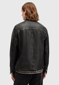 Giacca di pelle nera con superficie texturizzata, polsini con zip e vestibilità leggermente sagomata. Sotto è indossata una camicia a scacchi.