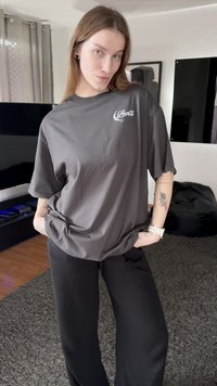 Gray cotton t-shirt with a relaxed fit, featuring a white logo on the chest, paired with black wide-leg pants. Plain background.