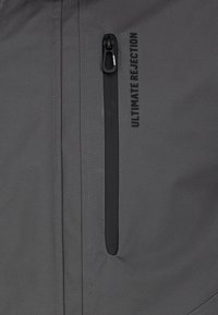 Gray jacket fabric with a smooth texture, featuring a zippered chest pocket and the text "ULTIMATE REJECTION" printed in black.
