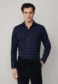 Camicia a quadri blu navy con controlli azzurri e marroni, colletto a punta e chiusura con bottoni, abbinata a pantaloni scuri.