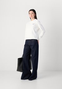 Kvinne står selvsikkert i en hvit genser og vide, mørke jeans, mens hun holder en svart tote-bag mot en lys bakgrunn.
