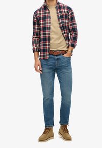 Man wearing blue jeans, tan suede boots, beige shirt, and unbuttoned red, white, and navy plaid shirt with brown belt and watch.