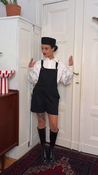 Femme avec un chapeau noir, une chemise blanche, une salopette courte noire et des bottes jusqu'aux genoux, debout à l'intérieur avec les bras levés près d'une porte blanche et d'un placard.