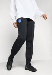 Sorte joggebukser med en blå Paris-logo-patch og en glidelåslomme, kombinert med en løs hvit hettegenser og sorte treningssko.