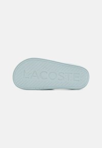 Suela de zapatilla de goma azul claro con estrías texturizadas y "LACOSTE" grabado de manera prominente en el centro. Acabado suave en general.