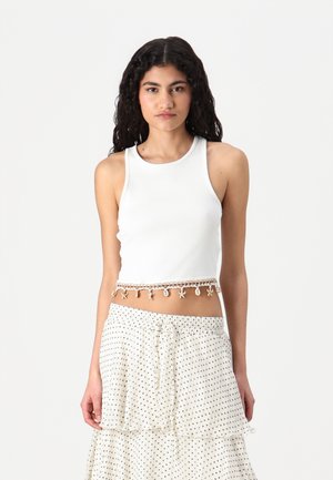 Débardeur blanc à côtes avec un design court et un ourlet décoré de perles. Le modèle le porte avec une jupe à pois.