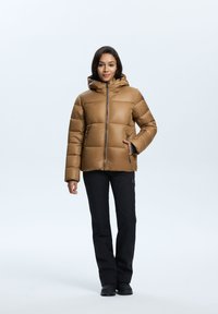 Doudoune marron avec capuche, fermeture éclair et poches latérales, présentant un design matelassé et des accents noirs. Portée avec un pantalon noir.