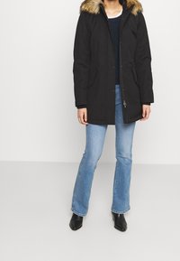 Svart parkas med en foderhuva av päls, har en framdragkedja och tryckknappar. Bärs över en blå tröja och ljusblå jeans.