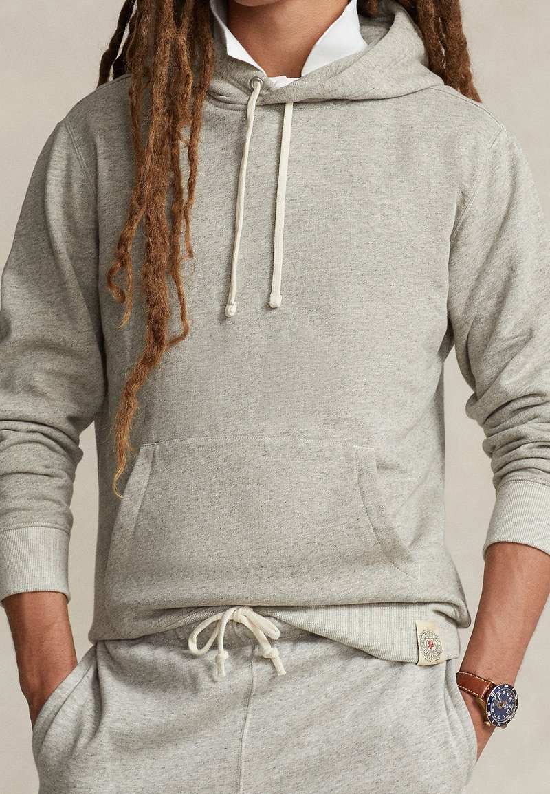 Personne avec de longues dreadlocks portant un sweat à capuche gris clair et un pantalon de jogging assorti, les mains dans les poches et une montre-bracelet en cuir marron.