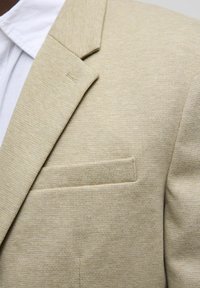 Light beige blazer with a textured fabric, featuring a notch lapel and a single chest pocket. Tailored fit with horizontal pattern detail.