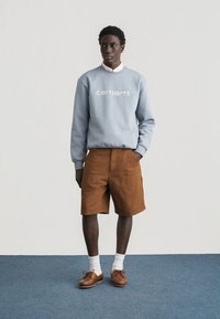 Άνδρας που φοράει γαλάζιο φούτερ Carhartt, καφέ σορτς, λευκές κάλτσες και καφέ δερμάτινα παπούτσια, στέκεται σε μπλε χαλί μπροστά σε λευκό τοίχο.