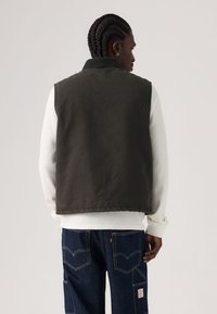 Σκούρο πράσινο αμάνικο γιλέκο με ψηλό γιακά, φορεμένο πάνω από λευκό μακρυμάνικο πουκάμισο. Διαθέτει λείο υλικό και απλό σχέδιο.