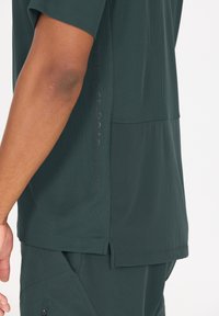 Korte mouwen donkergroene shirt van textuurmateriaal, met een netachtige patroon en zijsplitten. Heeft branding die langs de zijkant is gedrukt.