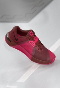 Red and pink athletic shoe with black Nike swoosh, mesh upper, and patterned sole on a light gray surface with white lines.