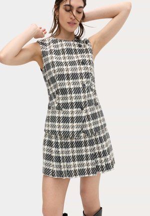 Vrouw in een mouwloze zwart-wit geruite jurk met knopen en zakken, poserend met opgeheven armen en gesloten ogen.