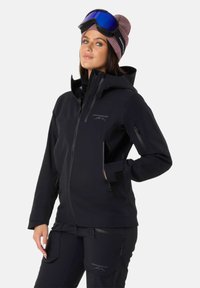 Giacca nera impermeabile con tasche con zip e cappuccio, abbinata a pantaloni neri con tasche cargo, accessoriata con un berretto rosa e occhiali.