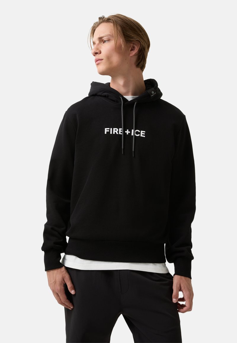 Jonge man in een zwarte "FIRE + ICE" hoodie en zwarte broek, kijkend naar de zijkant tegen een effen lichte achtergrond.