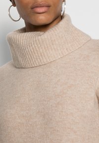 Pull à col roulé beige en tissu doux et texturé avec un col côtelé et des variations subtiles de couleur. Boucles d'oreilles argentées en forme de créoles visibles.