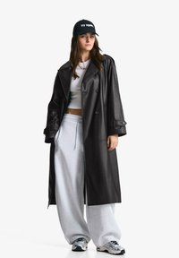 Svart läder trench coat med dubbelknäppta knappar, oversize passform, matchad med grå sweatpants och en svart keps med vit text.
