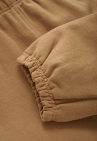 Close-up van camel-kleurige stof met details van een elastische manchet en zichtbare stiksels op zacht katoenmateriaal.